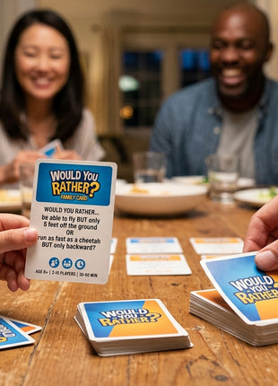 A close-up lifestyle photograph focusing on hands holding and drawing blue and yellow question cards from the "Would You Rather? Family Card Game" deck on a rustic wooden dining table. In the softly blurred background, a diverse family of four—parents and two kids—smiles and reacts to the gameplay under warm indoor lighting.