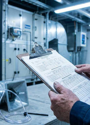 HVAC technician holding a clipboard and commissioning report while performing field diagnostics on a commercial air handling unit with digital air balancing equipment.