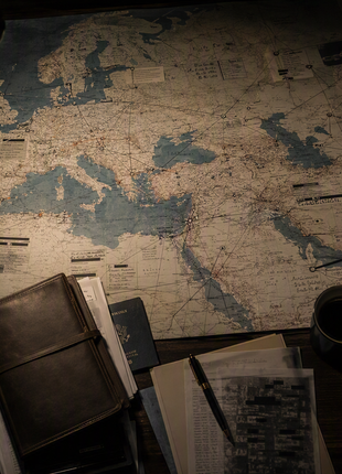 Professional intelligence-analysis workspace featuring a large international map of Europe, the Mediterranean, the Middle East, and Central Asia covered with strategic assessment markings, surrounded by intelligence reports, a leather notebook, passport, secure communications device, fountain pen, and coffee cup under cinematic low-key lighting.