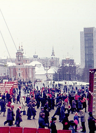 Vintage photograph of Moscow, USSR, showing crowds gathered in a city square during a Soviet celebration, surrounded by Cold War-era architecture, communist decorations, historic churches, and government buildings in 1977.