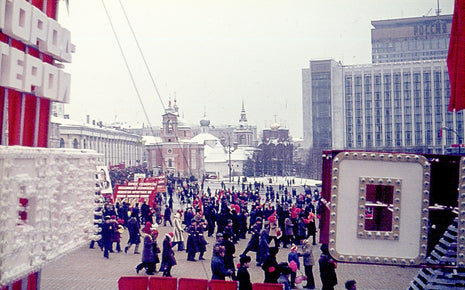 Vintage photograph of Moscow, USSR, showing crowds gathered in a city square during a Soviet celebration, surrounded by Cold War-era architecture, communist decorations, historic churches, and government buildings in 1977.