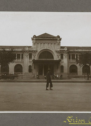 Historic architectural landmark in Saigon (now Ho Chi Minh City, Vietnam), featuring a grand French colonial-style public building with arched entrances, ornate façade details, and pedestrians crossing the street in front. This vintage photograph highlights the city's historic architecture during the French Indochina era and is in the public domain in the United States.