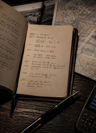 Close-up of an intelligence operative’s workspace featuring an open leather notebook with coded handwritten notes, a marked city map, passport, secure communications device, fountain pen, and operational documents illuminated by low-key desk lamp lighting in a cinematic espionage thriller setting.