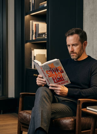 A reader sitting in a leather armchair in an modern apartment, engrossed in reading the hardcover spy novel The Silver Fish by Connor Martin.