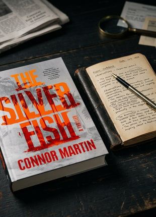Hardcover edition of The Silver Fish by Connor Martin placed next to a vintage leather reporter's notebook and metallic fountain pen on a dark wooden desk.