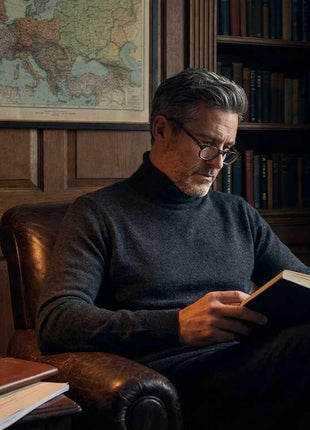 Man in a dark turtleneck and glasses reading a book in a leather chair, next to a desk lamp, a glass of whiskey, and an intelligence briefing document in a classic study.